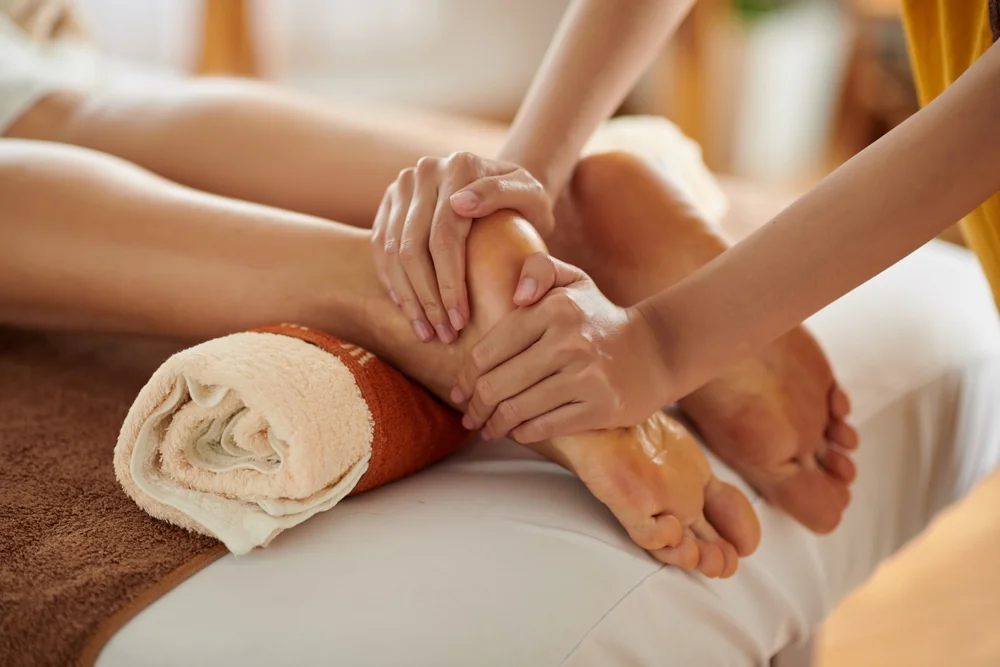 Therapist applying two-handed pressure across the top of a client's foot during foot reflexology, the ankle supported on a rolled terracotta towel